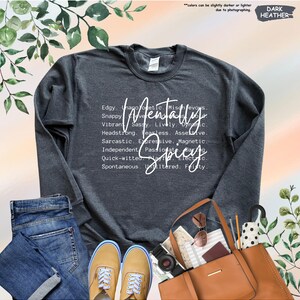 May include: Dark heather gray sweatshirt with the words "Mentally Spicy" in white script. The design includes a list of descriptive words. The sweatshirt is paired with jeans, sneakers, and a brown tote bag.