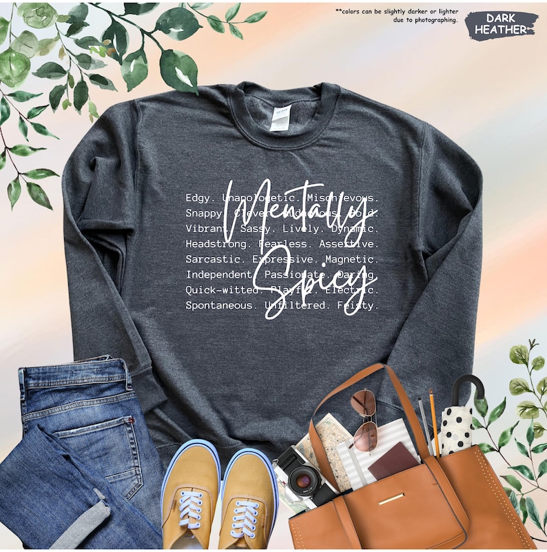 May include: Dark heather gray sweatshirt with the words "Mentally Spicy" in white script. The design includes a list of descriptive words. The sweatshirt is paired with jeans, sneakers, and a brown tote bag.