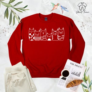 Puede incluir: Sudadera roja de manga larga con un gr&aacute;fico blanco de cinco gatos de dibujos animados. Los gatos son todos diferentes y tienen caracter&iacute;sticas &uacute;nicas, como lunares, rayas y diferentes expresiones faciales.