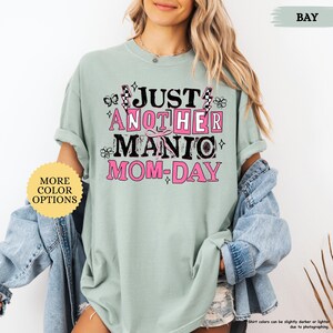 May include: A light green t-shirt with the text "JUST ANOTHER MANIC MOM-DAY" in pink and black. The shirt has rolled-up sleeves and a relaxed fit. A button with "MORE COLOR OPTIONS" is visible.