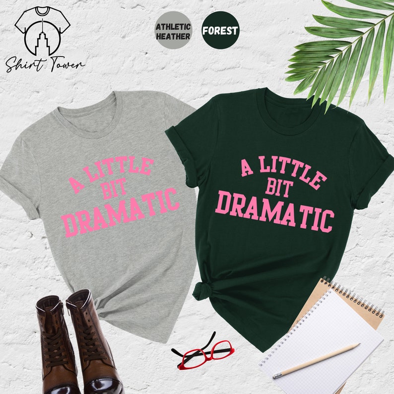 May include: Two t-shirts with the text "A Little Bit Dramatic" in pink lettering. The shirt on the left is a light grey colour and the shirt on the right is a dark green colour.