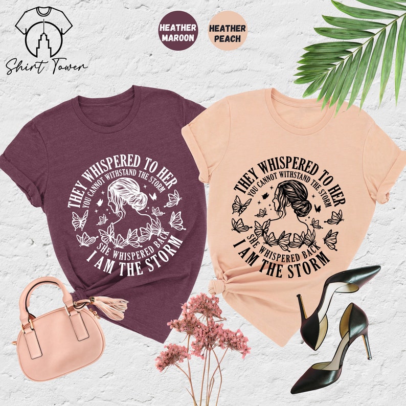May include: Two t-shirts with a graphic design featuring a woman with butterflies and the text "They whispered to her you cannot withstand the storm she whispered back I am the storm". The t-shirts are in heather maroon and heather peach.