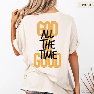 May include: Ivory-colored t-shirt with the text "GOD ALL THE TIME GOOD" in a combination of mustard yellow and black. The shirt's back is visible, with the text arranged vertically.
