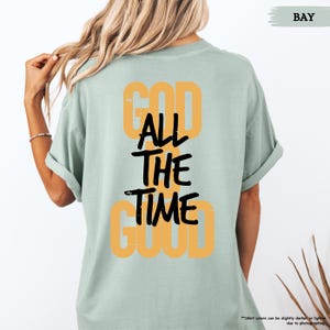 May include: A light green t-shirt with the text "GOD ALL THE TIME GOOD" in a combination of orange and black. The shirt has short sleeves and a relaxed fit. The word "BAY" is in the upper right corner.