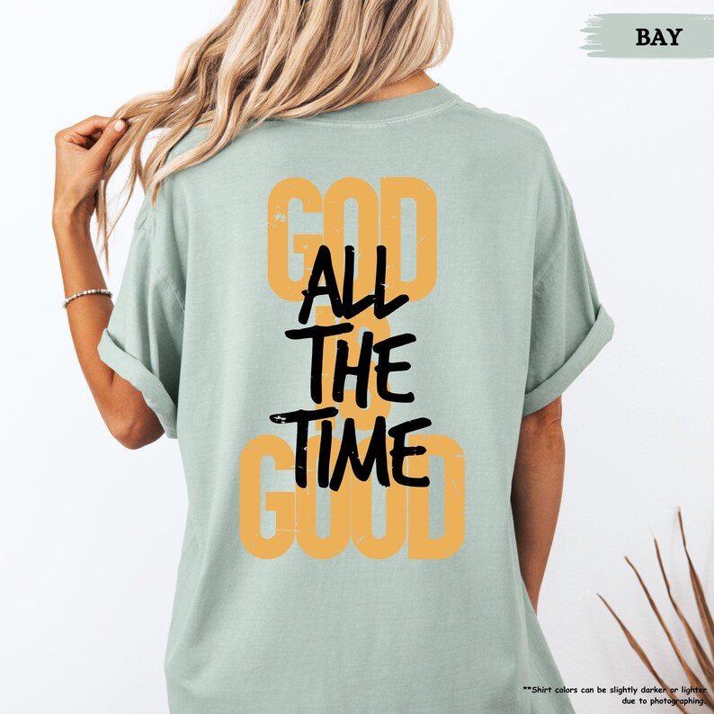 May include: A light green t-shirt with the text "GOD ALL THE TIME GOOD" in a combination of orange and black. The shirt has short sleeves and a relaxed fit. The word "BAY" is in the upper right corner.