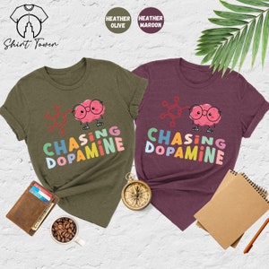 Pu&ograve; includere: Due t-shirt con la grafica di un cervello di cartone animato che indossa gli occhiali e una formula chimica. Il testo "Chasing Dopamine" &egrave; scritto in colori arcobaleno su entrambe le magliette. La maglietta a sinistra &egrave; di colore oliva melange e la maglietta a destra &egrave; di colore granata melange.