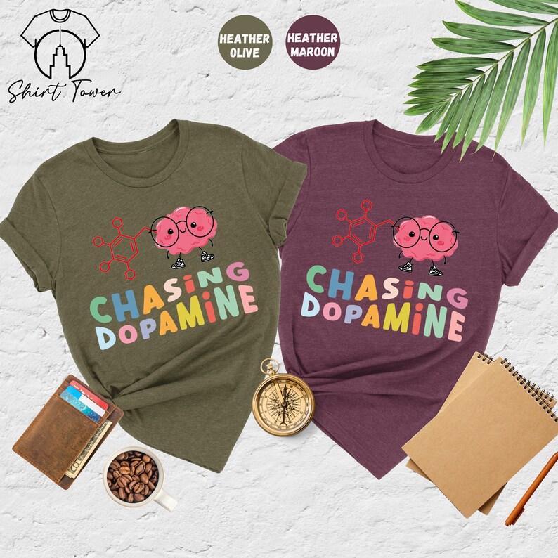 Pu&ograve; includere: Due t-shirt con la grafica di un cervello di cartone animato che indossa gli occhiali e una formula chimica. Il testo "Chasing Dopamine" &egrave; scritto in colori arcobaleno su entrambe le magliette. La maglietta a sinistra &egrave; di colore oliva melange e la maglietta a destra &egrave; di colore granata melange.