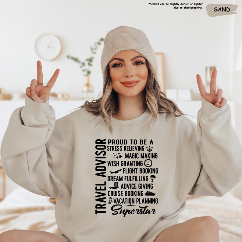 May include: A woman wearing a beige sweatshirt with the text "TRAVEL ADVISOR" and "PROUD TO BE A STRESS RELIEVING MAGIC MAKING WISH GRANTING FLIGHT BOOKING DREAM FULFILLING ADVICE GIVING CRUISE BOOKING VACATION PLANNING Superstar" printed on it.
