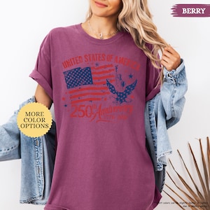 Op de afbeelding: Bessenkleurig T-shirt met een distressed afbeelding met de Amerikaanse vlag, het Vrijheidsbeeld en een adelaar. De tekst luidt "UNITED STATES OF AMERICA" en "250th Anniversary 1776-2026". Bevat de tekst "MORE COLOR OPTIONS".
