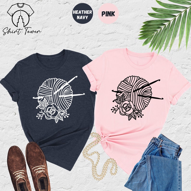 Pode incluir: Duas camisetas com um design de novelo de l&atilde; e agulhas de tric&ocirc;. A camiseta &agrave; esquerda &eacute; azul marinho mesclado e a camiseta &agrave; direita &eacute; rosa. O design &eacute; branco na camiseta azul marinho e preto na camiseta rosa.