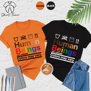 May include: Two t-shirts, one orange and one black, with the text "Human Beings 100% Organic Colors may vary." The text is in a rainbow of colors. The shirts are laid out on a table.