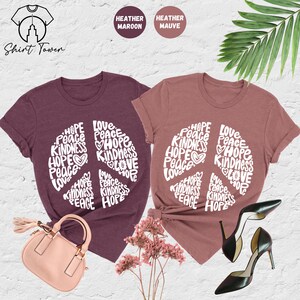 May include: Two heather colored t-shirts with a peace sign design made of the words "Hope", "Peace", "Kindness", and "Love". The shirts are available in heather maroon and heather mauve.
