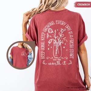 May include: A crimson red t-shirt with a white graphic design on the back. The design features a mason jar with flowers and the text "Keep going. Keep growing. Every day is a fresh start. You are & worth it."