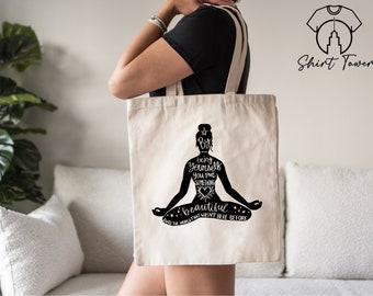 May include: A white canvas tote bag with black text and a silhouette of a person in a yoga pose. The text reads "By being yourself you put something beautiful into the world that wasn't here before".