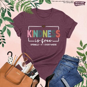 May include: A heather maroon t-shirt with the text "KINDNESS is free" in colorful letters with a distressed look. The text "SPRINKLE IT EVERYWHERE" is below the main text.