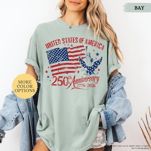 Op de afbeelding: Een lichtgroen t-shirt met een distressed afbeelding ter viering van de 250e verjaardag van de Verenigde Staten. Het ontwerp bevat de Amerikaanse vlag, het Vrijheidsbeeld en een adelaar. De tekst luidt "United States of America" en "250th Anniversary 1776-2026".