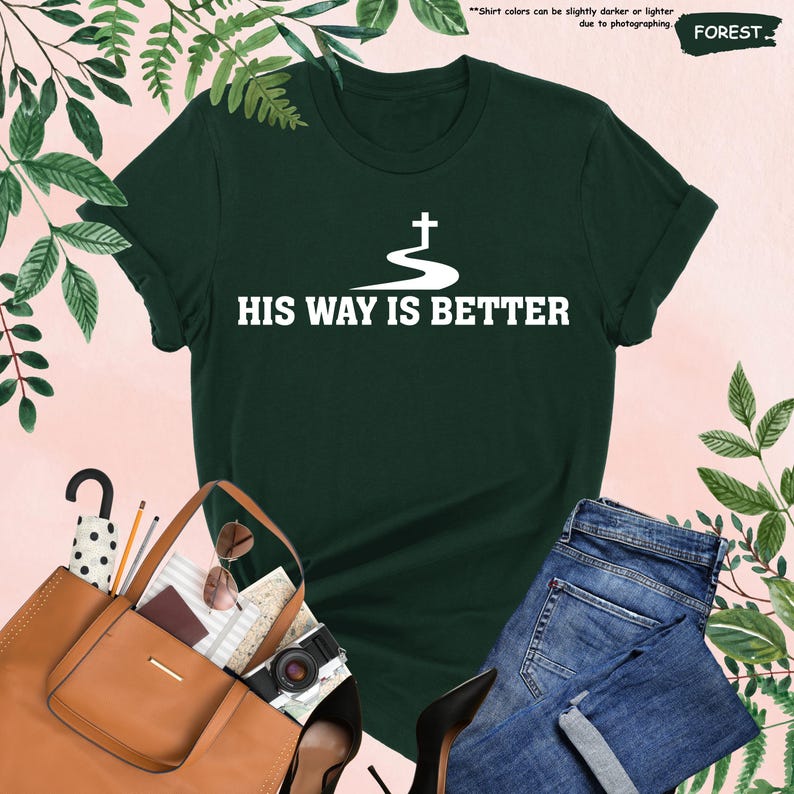 May include: A dark green t-shirt with the text "HIS WAY IS BETTER" in white. The graphic includes a cross and a flowing river design. The shirt is paired with blue jeans and a brown handbag.