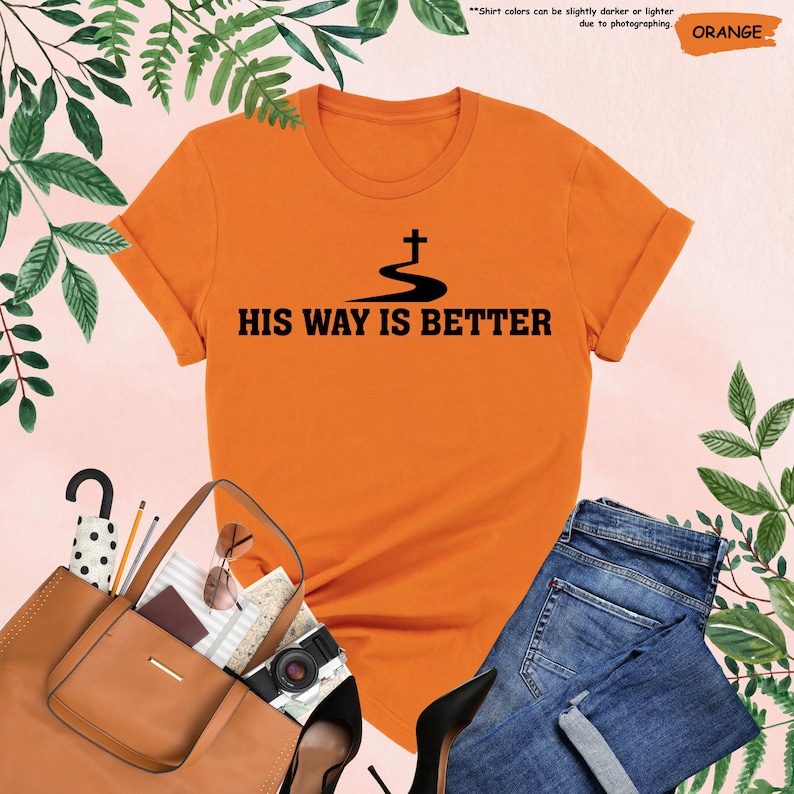 May include: An orange t-shirt with the text "HIS WAY IS BETTER" in black, featuring a cross design. The shirt is paired with blue jeans, a brown purse, and black heels. The image also includes a pair of sunglasses and a camera.