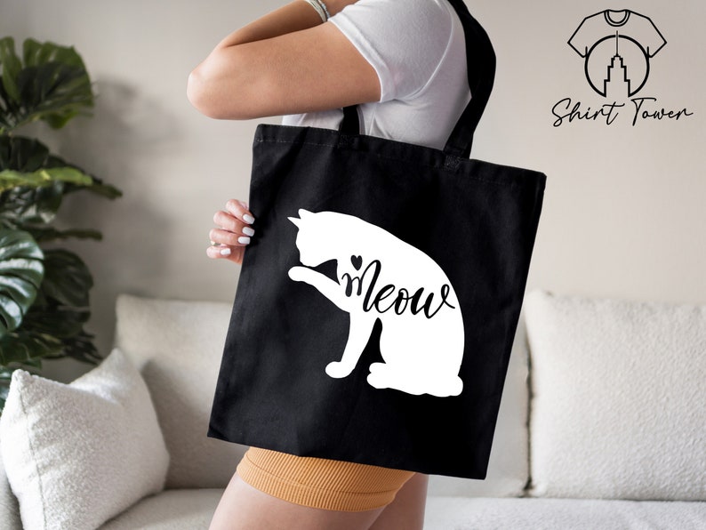Op de afbeelding: Een zwarte tote bag met een wit silhouet van een kat en het woord "Meow" in een cursief schrift.