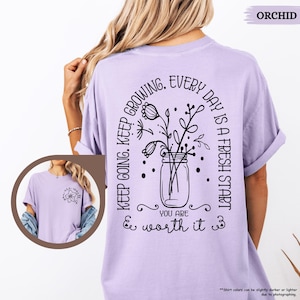 May include: A lavender t-shirt with a black graphic design featuring a jar of flowers and the text "Keep going. Keep growing. Every day is a fresh start. You are & worth it."