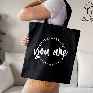 K&ouml;nnte beinhalten: Schwarze Einkaufstasche mit einem wei&szlig;en Kreisdesign, auf dem "You are loved, strong, important, worthy, beautiful" steht.
