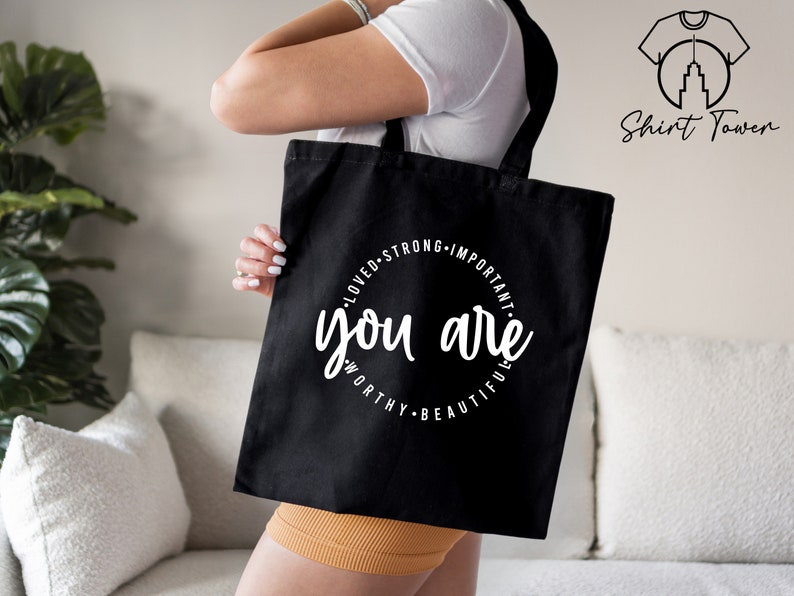 K&ouml;nnte beinhalten: Schwarze Einkaufstasche mit einem wei&szlig;en Kreisdesign, auf dem "You are loved, strong, important, worthy, beautiful" steht.