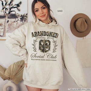 May include: A sand-colored hoodie with a front graphic that reads "ABANDONED Hunting Wives Social Club." The graphic includes a crest with a deer, guns, and a dove. The hoodie has a drawstring hood and a front pocket.