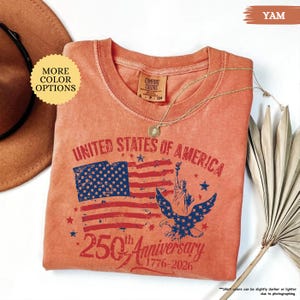 Op de afbeelding: Een yam-kleurig T-shirt met een afbeelding ter viering van de 250e verjaardag van de Verenigde Staten van Amerika. Het ontwerp omvat de Amerikaanse vlag, het Vrijheidsbeeld en een adelaar. Een gouden ketting ligt op het shirt.