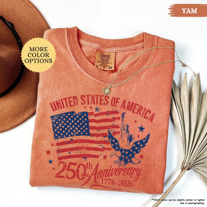 Op de afbeelding: Een yam-kleurig T-shirt met een afbeelding ter viering van de 250e verjaardag van de Verenigde Staten van Amerika. Het ontwerp omvat de Amerikaanse vlag, het Vrijheidsbeeld en een adelaar. Een gouden ketting ligt op het shirt.