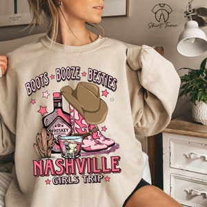 May include: A beige sweatshirt with a pink and brown graphic design. The graphic features a cowboy hat, boots, a bottle of whiskey, a glass of whiskey, and the text "Boots Booze Besties Nashville Girls Trip".