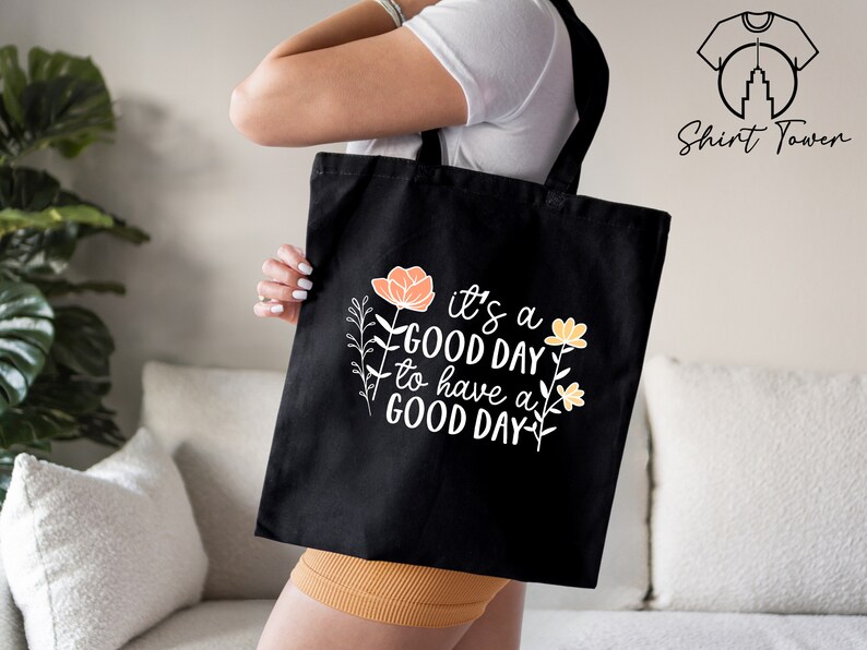 May include: Black tote bag with white text that reads "It's a good day to have a good day." The text is surrounded by orange and yellow flowers.