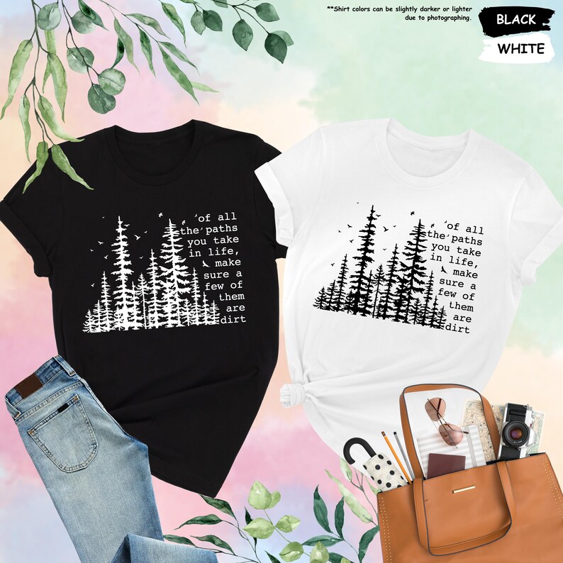 K&ouml;nnte beinhalten: Zwei T-Shirts mit einem schwarzen und wei&szlig;en Waldmotiv und dem Text 'of all the paths you take in life, make sure a few of them are dirt'. Die T-Shirts befinden sich auf einem rosa und wei&szlig;en Hintergrund.