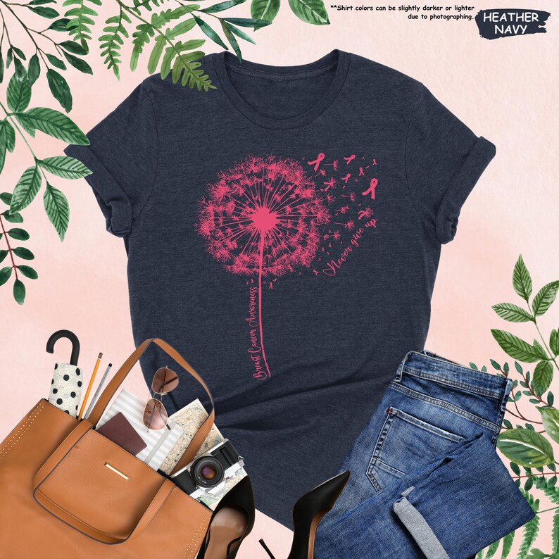 Puede incluir: Camiseta de color azul marino jaspeado con un dise&ntilde;o de diente de le&oacute;n rosa, lazos rosas y las palabras "Never give up" y "Breast Cancer Awareness". La camiseta se combina con vaqueros azules, un bolso tote marr&oacute;n y tacones negros.