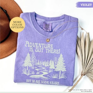 May include: A violet Comfort Colors t-shirt with the text "Adventure is Out There! But So Are Serial Killers" and a forest graphic. A gold necklace rests on the shirt. The shirt is a small size.