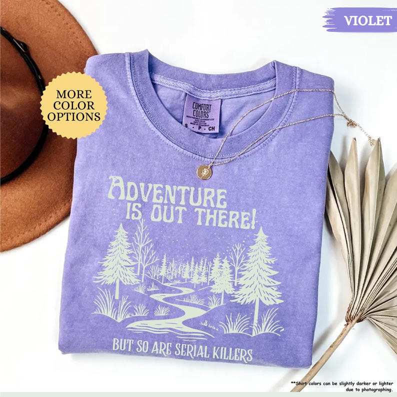 May include: A violet Comfort Colors t-shirt with the text "Adventure is Out There! But So Are Serial Killers" and a forest graphic. A gold necklace rests on the shirt. The shirt is a small size.
