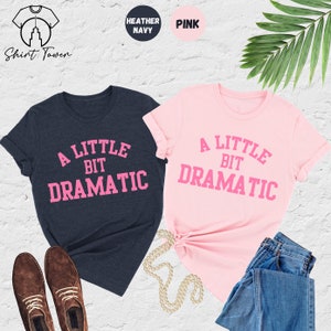 May include: Two t-shirts, one heather navy blue and one pink, with the text "A Little Bit Dramatic" in pink lettering.