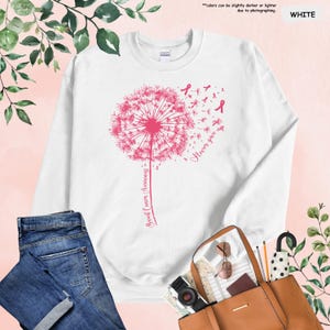 Puede incluir: Sudadera blanca con un dise&ntilde;o de diente de le&oacute;n rosa, lazos rosas y el texto "Breast Cancer Awareness" y "Never give up". La imagen incluye vaqueros azules y un bolso tote marr&oacute;n.