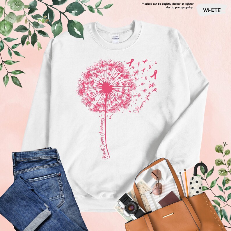 Puede incluir: Sudadera blanca con un dise&ntilde;o de diente de le&oacute;n rosa, lazos rosas y el texto "Breast Cancer Awareness" y "Never give up". La imagen incluye vaqueros azules y un bolso tote marr&oacute;n.