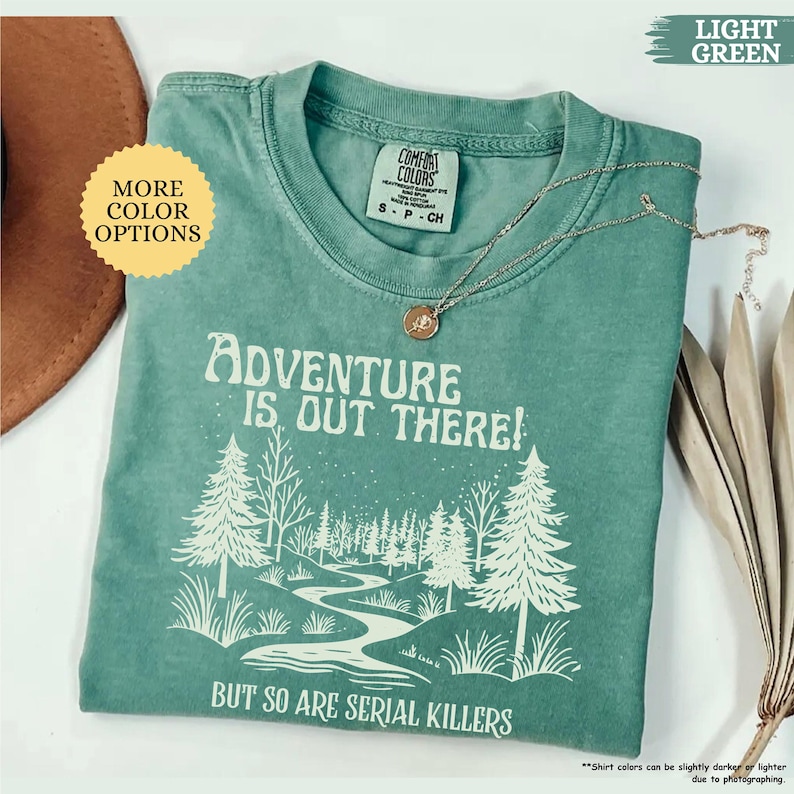 May include: A light green t-shirt with the text "ADVENTURE IS OUT THERE! BUT SO ARE SERIAL KILLERS" in white. The shirt features a forest graphic and a silver necklace with a pendant. The shirt is a Comfort Colors brand.