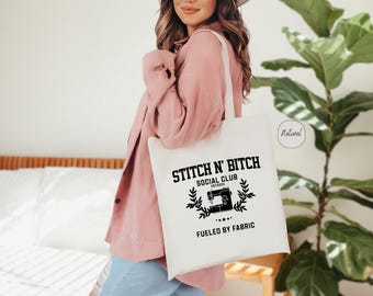 May include: White tote bag with black text that reads "Stitch 'n Bitch Social Club Established 2020 Fueled by Fabric". The text is surrounded by a black sewing machine graphic and a wreath of leaves.