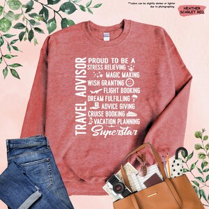 May include: A heather scarlet red sweatshirt with white text that reads "TRAVEL ADVISOR" and "PROUD TO BE A STRESS RELIEVING MAGIC MAKING WISH GRANTING FLIGHT BOOKING DREAM FULFILLING ADVICE GIVING CRUISE BOOKING VACATION PLANNING SUPERSTAR".