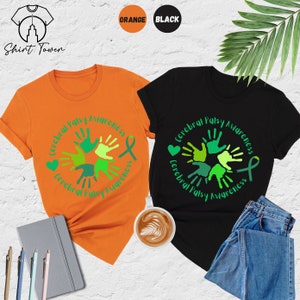 May include: Two t-shirts, one orange and one black, with a design of green handprints in a circle and the text "Cerebral Palsy Awareness".