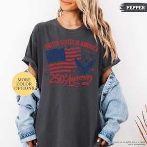 Op de afbeelding: Donkergrijs t-shirt met een verweerde Amerikaanse vlag en de tekst "UNITED STATES OF AMERICA" en "250th Anniversary 1776-2026". Het shirt heeft opgerolde mouwen en een losse pasvorm. Het shirt is gemarkeerd met "PEPPER" en toont "MORE COLOR OPTIONS".