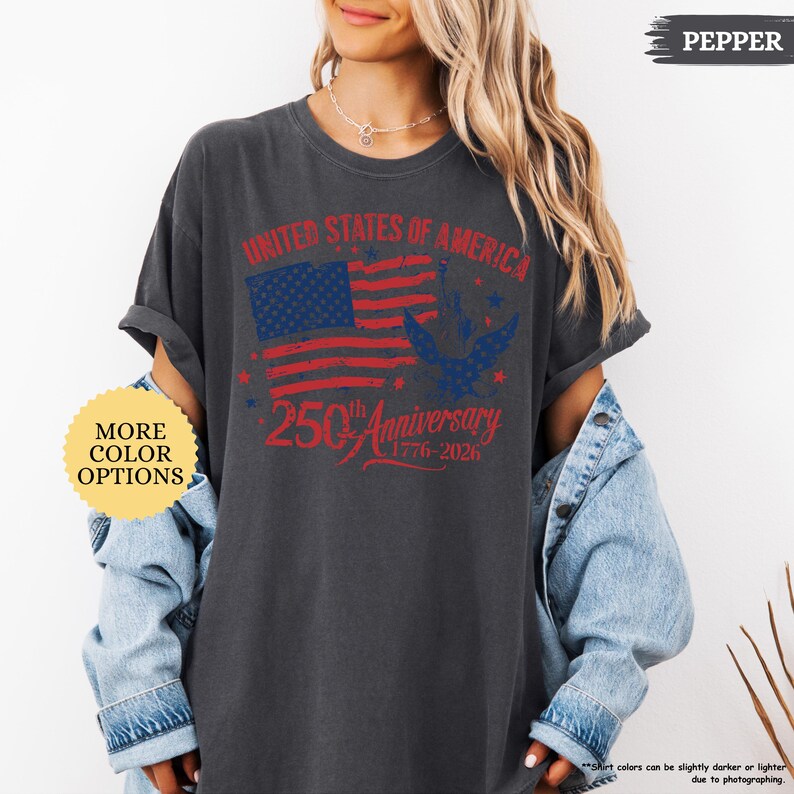 Op de afbeelding: Donkergrijs t-shirt met een verweerde Amerikaanse vlag en de tekst "UNITED STATES OF AMERICA" en "250th Anniversary 1776-2026". Het shirt heeft opgerolde mouwen en een losse pasvorm. Het shirt is gemarkeerd met "PEPPER" en toont "MORE COLOR OPTIONS".