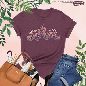 Op de afbeelding: Bordeauxrood heather T-shirt met de tekst "Healing is Not Linear" in een golvend ontwerp met regenboogkleuren. Het shirt wordt getoond met een bruine tote bag, zonnebril, camera en een spijkerbroek.