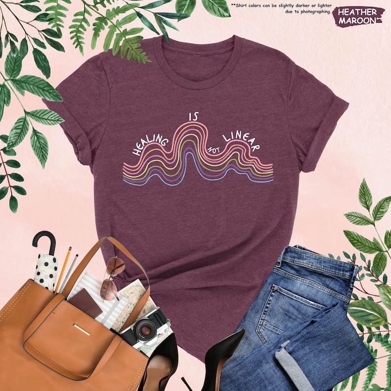 Op de afbeelding: Bordeauxrood heather T-shirt met de tekst "Healing is Not Linear" in een golvend ontwerp met regenboogkleuren. Het shirt wordt getoond met een bruine tote bag, zonnebril, camera en een spijkerbroek.