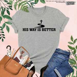 May include: Athletic heather gray t-shirt with the text "HIS WAY IS BETTER" in black. The shirt features a black graphic of a cross above a winding path. The shirt is paired with blue jeans, a brown purse, and black heels.