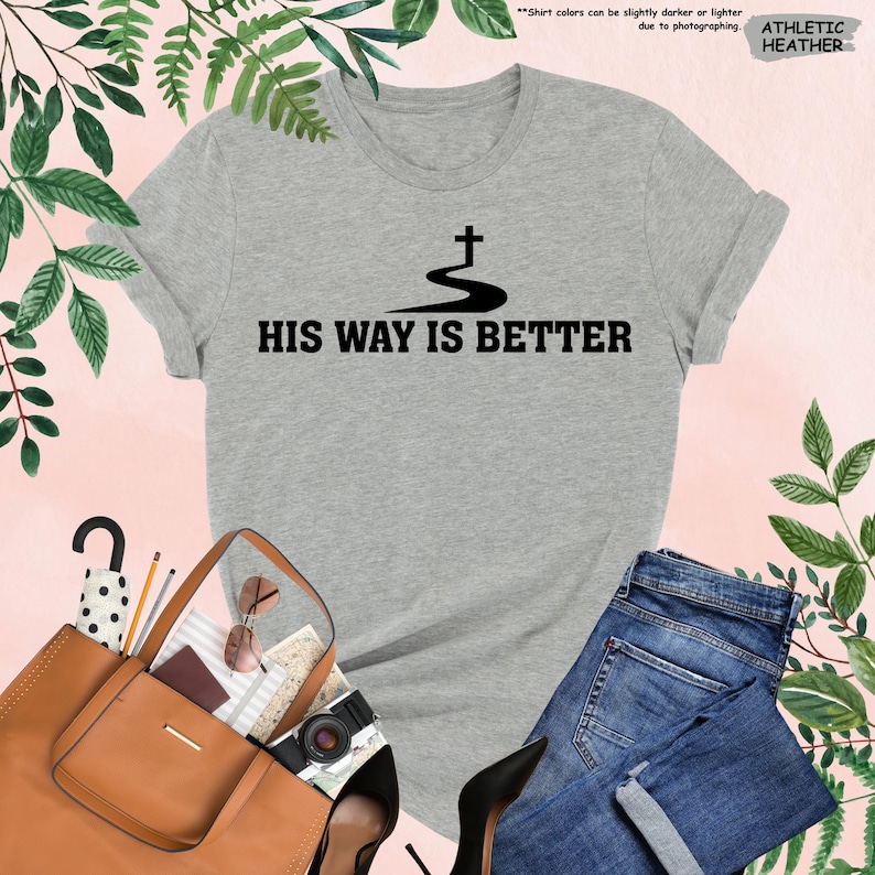 May include: Athletic heather gray t-shirt with the text "HIS WAY IS BETTER" in black. The shirt features a black graphic of a cross above a winding path. The shirt is paired with blue jeans, a brown purse, and black heels.