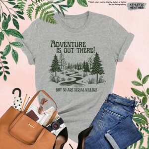 Op de afbeelding: Athletic heather grijs t-shirt met de tekst "Adventure is Out There! But So Are Serial Killers" en een bosafbeelding. Het shirt wordt gecombineerd met een blauwe jeans, een bruine handtas en zwarte hakken.