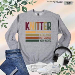 May include: A gray sweatshirt with the word "Knitter" in a vintage style font. The text is layered with colorful stripes and the words "Yarn Magician", "Needle Ninja", "Cozy Creator", and "Wool Wizard" are written below.